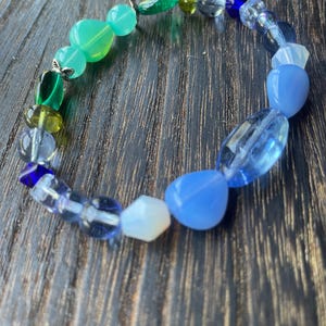 May include: A beaded bracelet featuring a variety of colorful beads. The bracelet includes green, blue, and clear beads in various shapes and sizes. The beads are strung together on a clear elastic cord, creating a unique accessory.