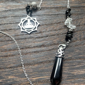 May include: A silver-colored pendulum with a chain, featuring a star-shaped pendant with a seated figure, black and clear crystal beads, and a black, pointed crystal. The pendulum hangs against a dark wood grain background.