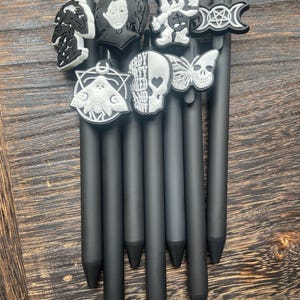 May include: A collection of black pens with decorative toppers. The toppers feature various designs, including a skull, a butterfly, a moth, and a cartoon character. Some toppers are black and white, while others have additional colors. The pens are arranged on a wooden surface.