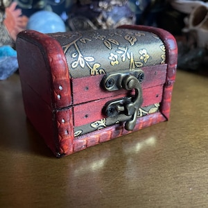 Puede incluir: Un pequeño cofre de madera rectangular con tapa abisagrada. El cofre está teñido de rojo con un patrón floral decorativo en la tapa y los lados. Cuenta con un cierre y bisagras de metal.