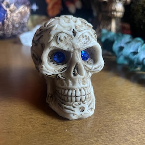 May include: A detailed, off-white skull figurine with blue gemstone eyes. The skull has intricate carvings and a detailed jawline. The background includes other decorative items, suggesting a gothic or Halloween theme.