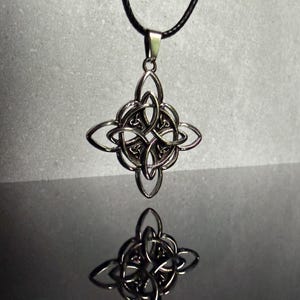 May include: A silver-toned metal pendant featuring an intricate, symmetrical four-pointed knot design with interlaced loops. The pendant is suspended from a thin black cord and displays a clear reflection on the surface beneath it.
