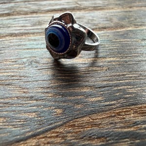 Puede incluir: Anillo plateado con un diseño central de ojo azul y negro. El ojo está rodeado por un engaste texturizado en forma de flor. El anillo se presenta sobre un fondo de madera, resaltando los detalles de la joya.