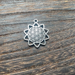 May include: Silver-colored pendant with a flower of life design. The pendant has a circular shape with a detailed geometric pattern inside, surrounded by pointed petals. The pendant is shown on a dark wooden surface.