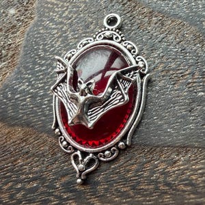 May include: Silver-toned pendant with a red oval center and a bat design. The bat is in the center of the pendant with its wings spread. The pendant has an ornate, filigree-style frame and a small loop at the top for a chain.