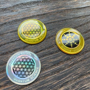 May include: Three circular Quantum Science tokens with a geometric pattern. The tokens have a gold or silver metallic finish with the words "Quantum Shield" and "Quantum Science" printed around the edge. The background is a dark wooden surface.