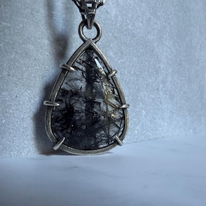 May include: A teardrop-shaped rutilated quartz pendant featuring black needle-like inclusions encased in a silver-toned metal setting with four prongs. The pendant hangs from a matching silver-toned chain with rectangular links.