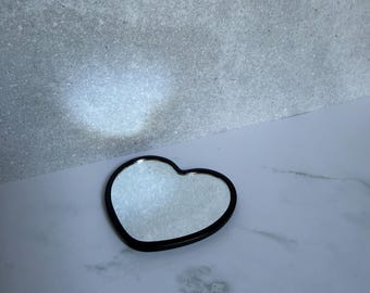 Op de afbeelding: Een hartvormige spiegel met een gladde, effen zwarte rand. Het reflecterende oppervlak is helder en heeft een hartvormig silhouet, wat een duidelijk contrast vormt met de donkere rand die de gehele vorm van het object omlijnt.
