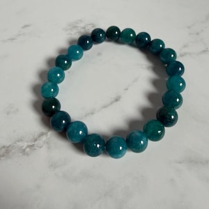 May include: A circular stretch bracelet composed of polished, spherical beads in varying shades of deep teal, forest green, and ocean blue. The beads feature a smooth, glossy finish with subtle internal variations in color and translucency throughout.