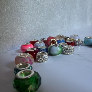 May include: A collection of assorted jewelry beads featuring various colors, patterns, and textures. The beads include faceted green glass, pink floral designs, clear rhinestones, and solid colored spheres, all with silver-toned metal cores.