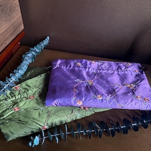Puede incluir: Dos pequeñas bolsas de satén bordadas. Una es verde con un diseño floral y la otra es morada con un diseño floral.