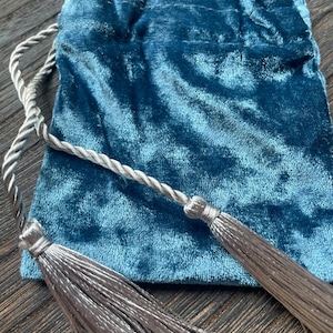 Puede incluir: Una bolsa de terciopelo color turquesa con cordón y cierre de borlas gris plateado. La bolsa tiene un aspecto suave y texturizado. Las borlas son largas y detalladas.