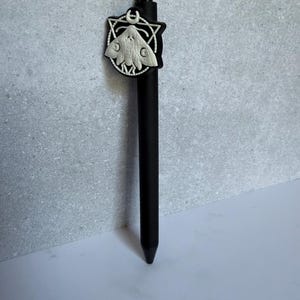 May include: A matte black retractable ballpoint pen featuring a circular topper with a white embossed moth design centered within a geometric frame and a small crescent moon symbol positioned at the top of the circular emblem.