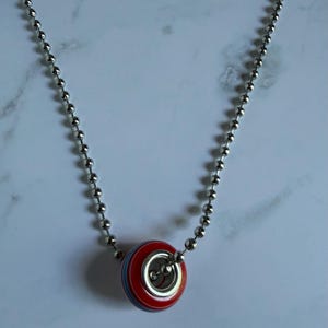 May include: A silver-toned ball chain necklace featuring a single round bead pendant. The bead is primarily red with thin horizontal stripes in light blue and white, and it has a metallic silver-coloured centre insert through the middle.