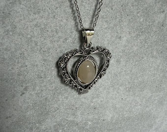 Op de afbeelding: Een zilverkleurige ketting met een hartvormige hanger met een gedetailleerde filigraanrand. In het midden van het hart bevindt zich een ovale, gladde, lichtbeige cabochonsteen. De hanger hangt aan een fijne zilverkleurige metalen ketting.