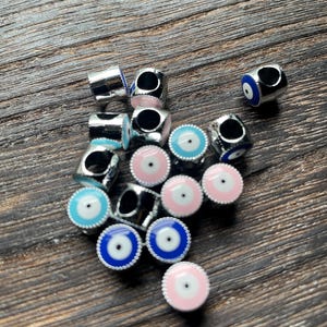 May include: A collection of silver-toned metal beads with an eye design. The beads feature a central black pupil surrounded by blue or pink concentric circles. The beads are scattered on a dark wood surface.