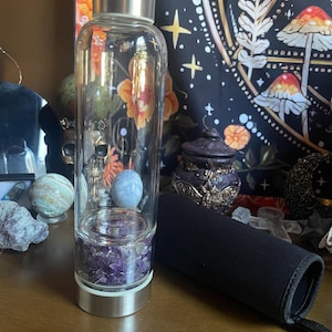 Puede incluir: Una botella de agua de vidrio transparente con tapa y base plateadas. La botella contiene cristales de amatista en la parte inferior, con una piedra azul y verde en el interior. Una funda de neopreno negra descansa junto a la botella.