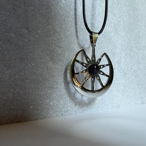 May include: A silver-toned metal pendant featuring a crescent moon shape integrated with a sunburst design. A small, dark, speckled circular stone is set in the center. The pendant hangs from a thin black cord necklace against a neutral background.