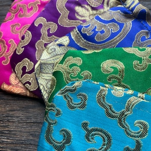 Puede incluir: Primer plano de cuatro bolsas de tela coloridas. Las bolsas son en tonos rosa, morado, azul, verde y turquesa. Cada bolsa presenta un patrón dorado ornamentado. Las bolsas tienen forma redondeada y están hechas de un material brillante y texturizado.