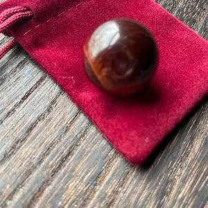 Könnte beinhalten: Eine polierte, braune Edelstein-Kugel liegt auf einem kleinen, tiefroten Samtbeutel. Der Edelstein hat eine glänzende Oberfläche, die das Licht reflektiert. Der Beutel hat einen Kordelzugverschluss. Der Hintergrund ist eine dunkle Holzmaserung.