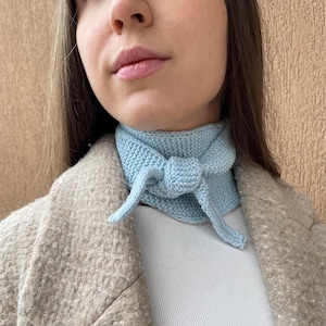 May include: A light blue knitted scarf tied in a bow around the neck. The scarf is made of a soft, textured yarn.
