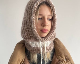 May include: A knitted hooded neck warmer in a light brown and white color scheme. The neck warmer has a drawstring and is made of a soft, textured yarn.