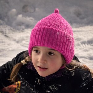 Op de afbeelding: Een felroze gebreide beanie-muts met een klein detail aan de bovenkant. De muts wordt gedragen door een persoon die een zwarte jas draagt, met een besneeuwde achtergrond.