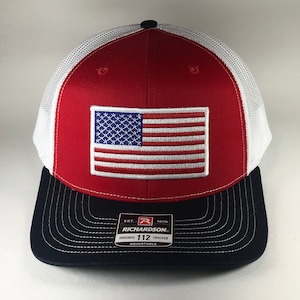 May include: Red, white, and blue trucker hat with an American flag patch on the front. The hat has a black mesh back and a black brim with the text "Richardson 112 Original Trucker Adjustable" on the back.