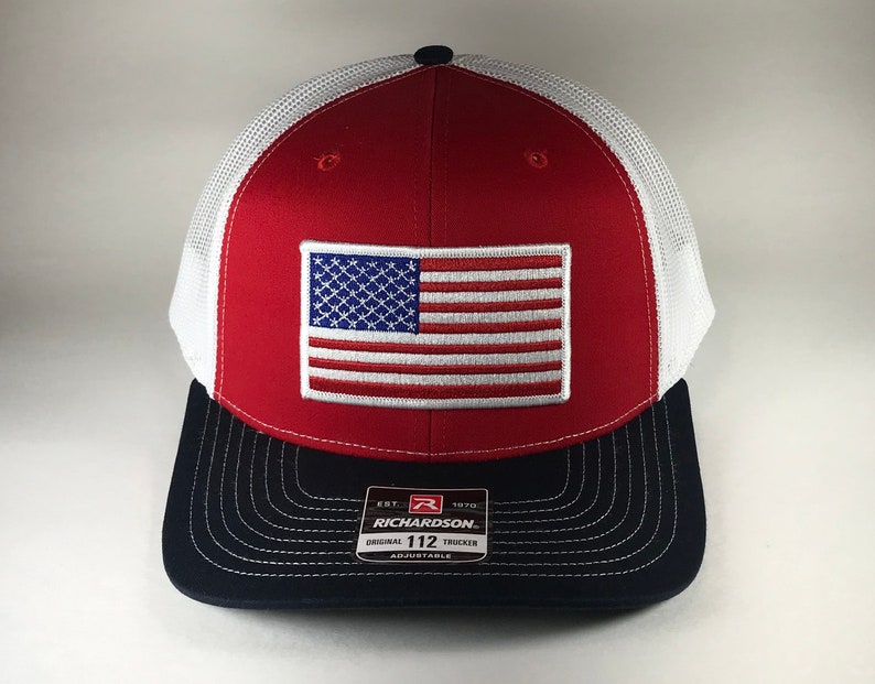 May include: Red, white, and blue trucker hat with an American flag patch on the front. The hat has a black mesh back and a black brim with the text "Richardson 112 Original Trucker Adjustable" on the back.
