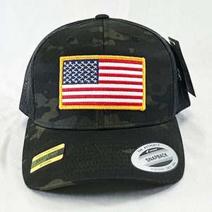 May include: A black and green camouflage trucker hat with a red, white, and blue American flag patch on the front. The hat has a black mesh back and a black adjustable snap closure. The text "THE AUTHENTIC SNAPBACK" is printed on a silver tag on the back of the hat.