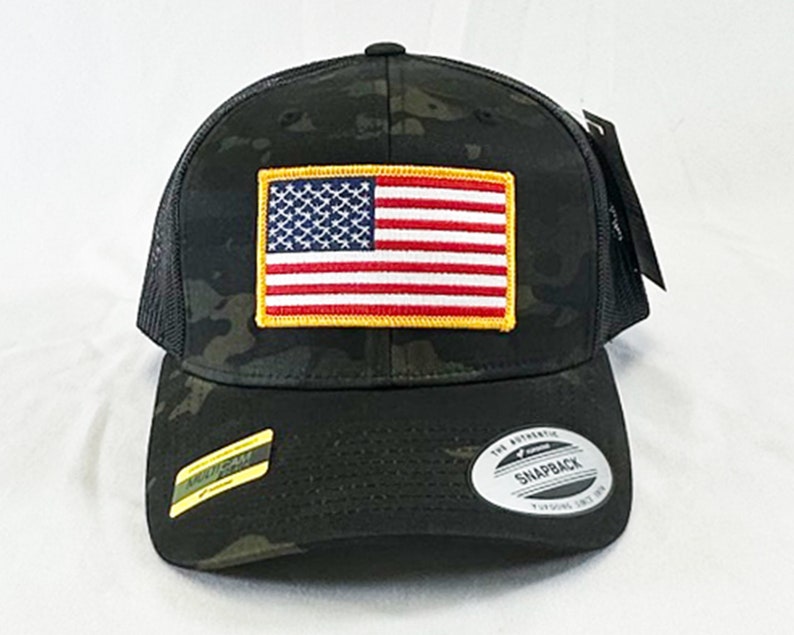 May include: A black and green camouflage trucker hat with a red, white, and blue American flag patch on the front. The hat has a black mesh back and a black adjustable snap closure. The text "THE AUTHENTIC SNAPBACK" is printed on a silver tag on the back of the hat.