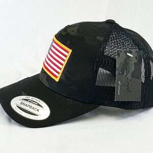 May include: Black and green camouflage trucker hat with a black mesh back. The hat has a small American flag patch on the front and a black tag with the text "AUTHENTIC" on the side.