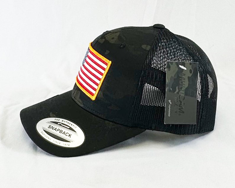 May include: Black and green camouflage trucker hat with a black mesh back. The hat has a small American flag patch on the front and a black tag with the text "AUTHENTIC" on the side.