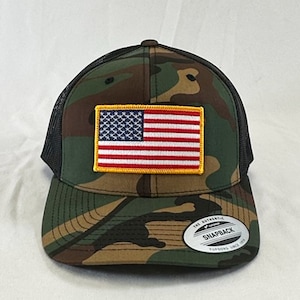 May include: A black and green camouflage trucker hat with a red, white, and blue American flag patch on the front. The hat has a black mesh back and a black brim. The hat has a white label on the back that says "The Authentic Snapback Yupoong Since 1975."