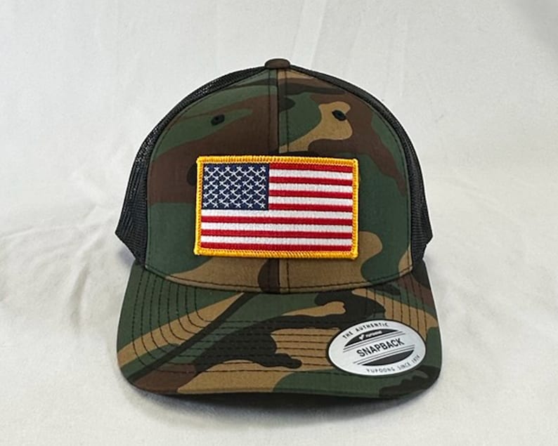 May include: A black and green camouflage trucker hat with a red, white, and blue American flag patch on the front. The hat has a black mesh back and a black brim. The hat has a white label on the back that says "The Authentic Snapback Yupoong Since 1975."