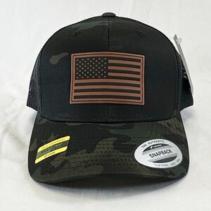 May include: A black and green camouflage trucker hat with a brown leather American flag patch on the front. The hat has a mesh back and a snap closure. The hat has a "Snapback" label on the back.