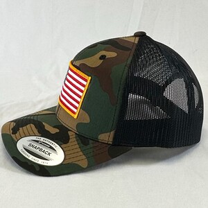 May include: A camouflage trucker hat with a black mesh back and a small American flag patch on the front. The hat has a sticker on the side that says "The Authentic Snapback, Fitted Since 1974."