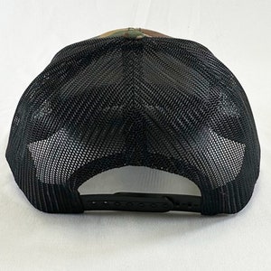 May include: A black and green camouflage trucker hat with a black mesh back. The hat has a black adjustable strap.