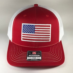 May include: Red and white trucker hat with a white mesh back. The hat features an embroidered American flag patch on the front and the text "EST. 1970 RICHARDSON. ORIGINAL 112 TRUCKER ADJUSTABLE" on the back.