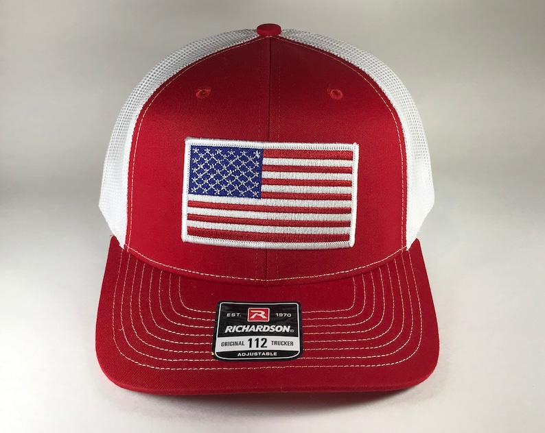 May include: Red and white trucker hat with a white mesh back. The hat features an embroidered American flag patch on the front and the text "EST. 1970 RICHARDSON. ORIGINAL 112 TRUCKER ADJUSTABLE" on the back.