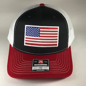 May include: Black and white trucker hat with a red bill. The hat features a white mesh back and a red, white, and blue American flag patch on the front. The hat also has a black label with the text "Richardson 112 Original Trucker Adjustable" on the front.