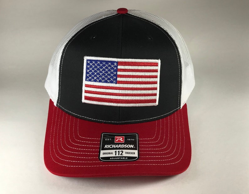 May include: Black and white trucker hat with a red bill. The hat features a white mesh back and a red, white, and blue American flag patch on the front. The hat also has a black label with the text "Richardson 112 Original Trucker Adjustable" on the front.
