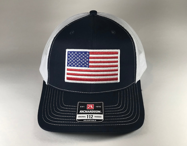 May include: A navy blue and white trucker hat with an embroidered American flag patch on the front. The hat has a white mesh back and an adjustable strap with the Richardson 112 logo.