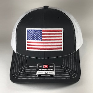 May include: Black and white trucker hat with a red, white, and blue American flag patch on the front. The hat has a white mesh back and a black brim. The hat has a label on the side that says "Richardson 112 Original Trucker Adjustable".