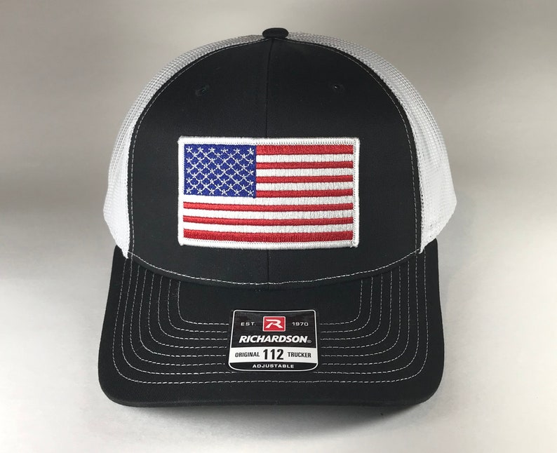 May include: Black and white trucker hat with a red, white, and blue American flag patch on the front. The hat has a white mesh back and a black brim. The hat has a label on the side that says "Richardson 112 Original Trucker Adjustable".