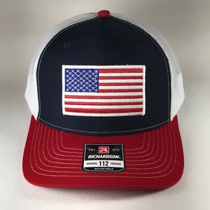 May include: A navy blue and white trucker hat with a red, white, and blue American flag patch on the front. The hat has a red undervisor and a white mesh back. The hat has a black label on the back that says "Richardson Est. 1970 Original 112 Trucker Adjustable".