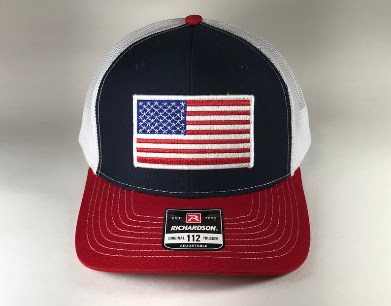 May include: A navy blue and white trucker hat with a red, white, and blue American flag patch on the front. The hat has a red undervisor and a white mesh back. The hat has a black label on the back that says "Richardson Est. 1970 Original 112 Trucker Adjustable".