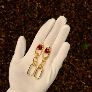 May include: A pair of gold chain earrings with red gemstone accents. The earrings are dangling and feature a chain link design.