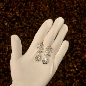 May include: A pair of silver dangle earrings with clear crystal stones. The earrings are displayed on a white glove.