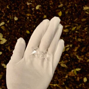 May include: Two clear crystal star-shaped earrings on a white glove against a background of brown leaves.
